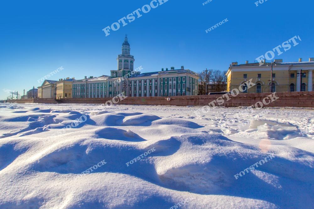 На фотографии изображена Кунсткамера — одно из старейших зданий Санкт-Петербурга, расположенное на Университетской набережной. Передний план покрыт снежными сугробами, создающими ощущение зимней тишины и спокойствия. Яркое голубое небо подчёркивает архитектурную красоту здания. Это место является символом исторического и культурного наследия города.